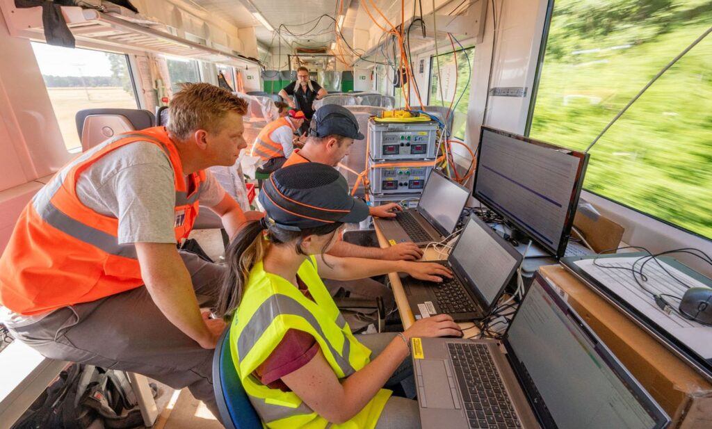 Mehrere Personen in Warnwesten sitzen in einem fahrenden Zug an einer Teststation, die aus Laptops und Monitoren besteht.