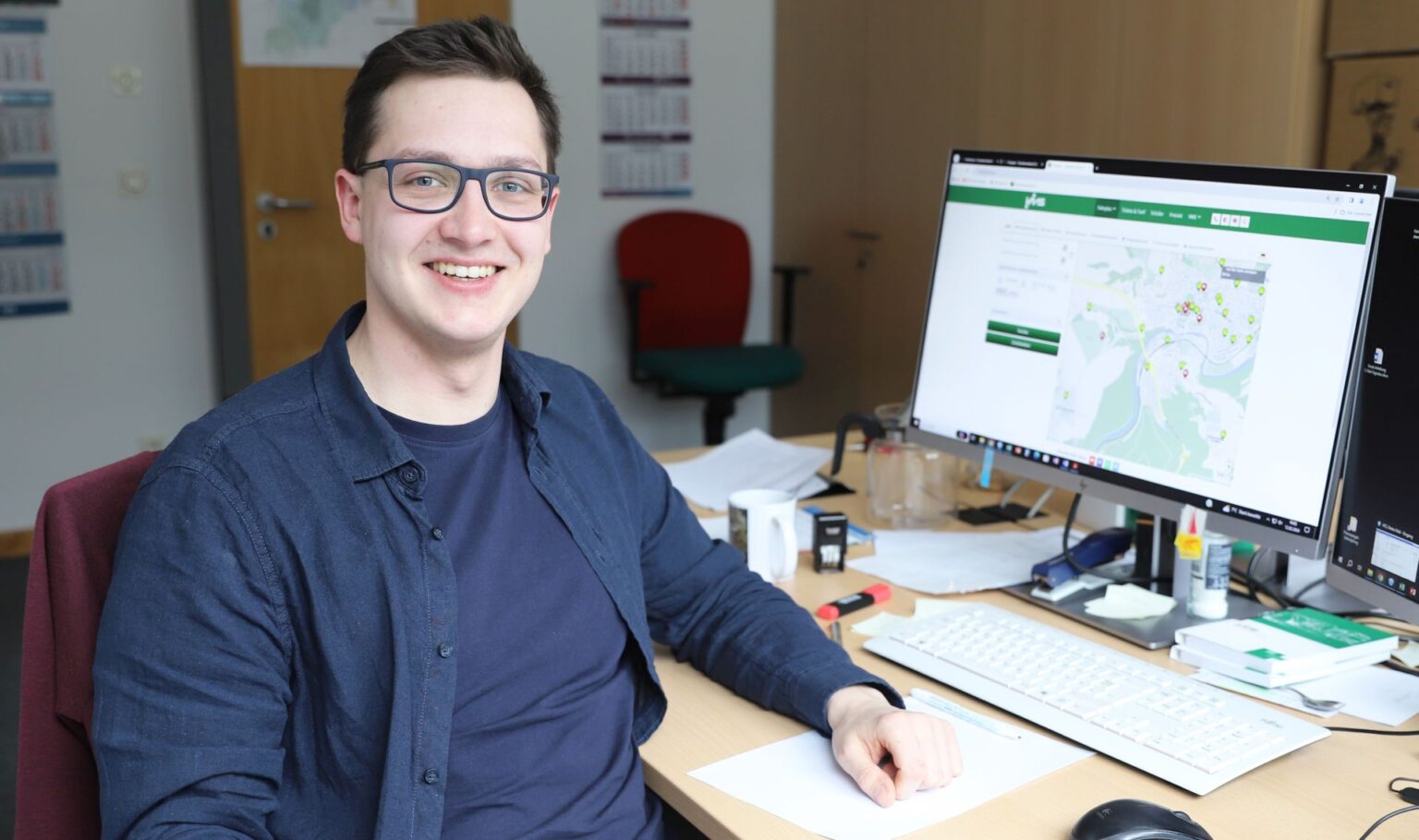 Ein junger Mann sitzt am Schreibtisch und lächelt in die Kamera.