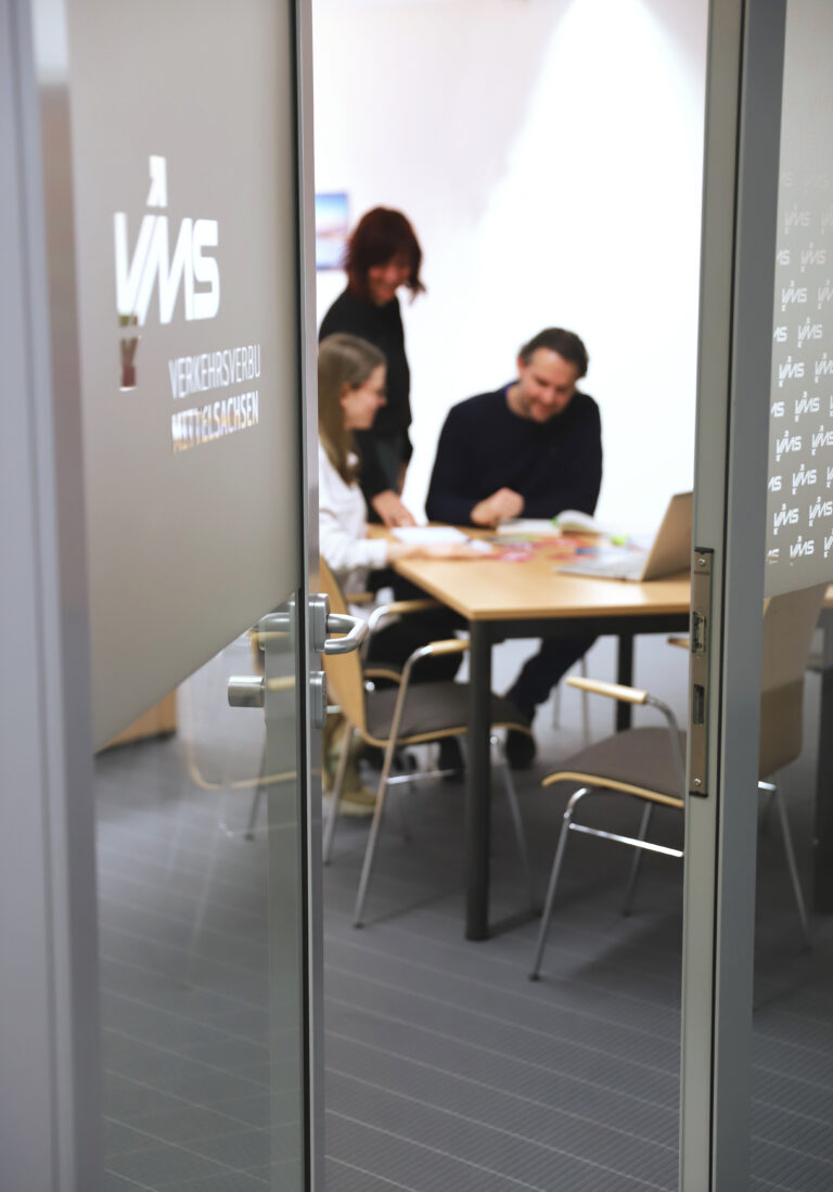 Mehrere Personen sitzen an einem Tisch und schauen auf Dokumente, das Foto wurde durch eine halb geöffnete Tür geschossen, auf der "VMS" steht.