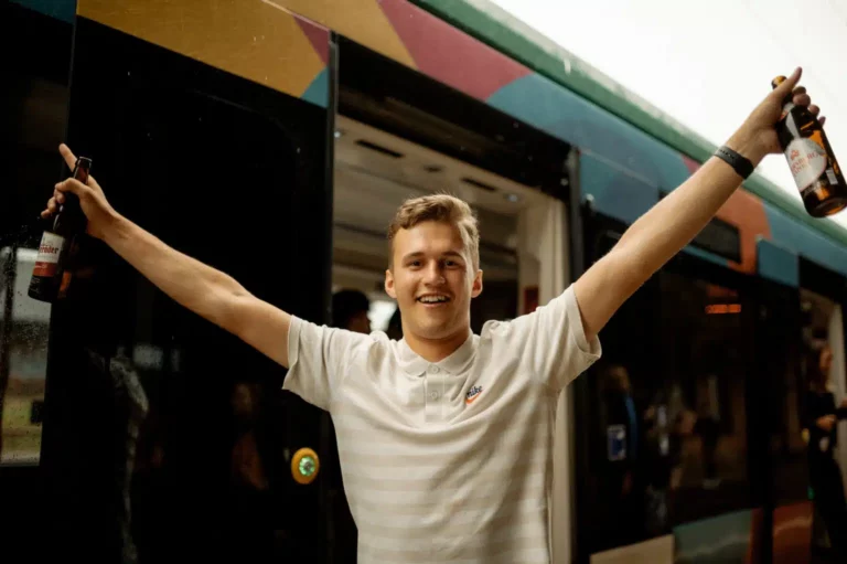 Ein froher junger Mann steht mit erhobenen Armen vor der Tür einer Straßenbahn.
