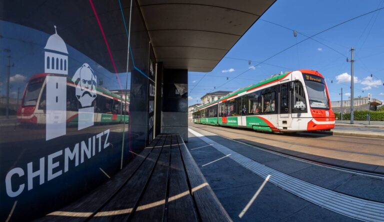 Eine City-Bahn fährt über den Bahnhofsvorplatz, rechts ist auf einem Schaufenster das Wort "Chemnitz" sowie ein Piktogramm des roten Turms und des Karl-Marx-Kopfes zu sehen.