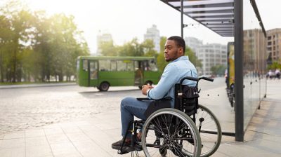 Young disabled black man in wheelchair waiting for public transport on bus stop, having difficulty traveling around city, copy space. Millennial handicapped guy at urban auto station